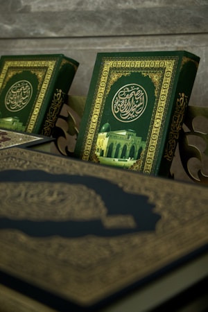 A close-up view of ornate books with intricate Arabic calligraphy and a picture of a mosque on their covers. The book covers feature a rich green background with gold embellishments.