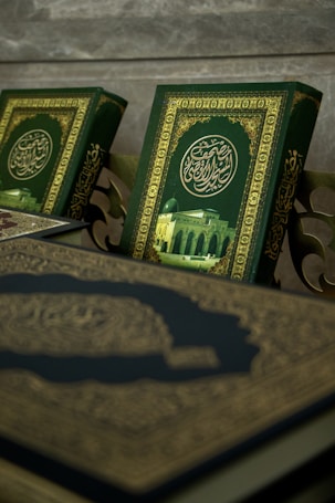 A close-up view of ornate books with intricate Arabic calligraphy and a picture of a mosque on their covers. The book covers feature a rich green background with gold embellishments.