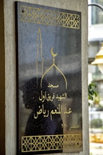A black plaque with gold Arabic script, featuring a mosque design with a minaret and crescent moon. Decorative gold patterns adorn the edges. The reflection of a person's silhouette is visible against the shiny surface. The background includes part of an outdoor scene with a lamp post.