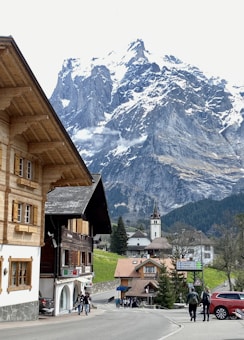 A charming alpine village with traditional wooden buildings, surrounded by towering snow-capped mountains. A church with a tall steeple is centrally located among the houses. A few people can be seen walking along the street, and there are green fields and dense forests in the background.
