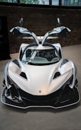 A sleek, futuristic sports car with gullwing doors raised. The silver vehicle has an aerodynamic design emphasizing speed and performance, with sharp lines and distinctive features. The car is situated indoors against a concrete background, with some reflections visible in its shiny surface.