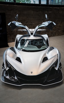 A sleek, futuristic sports car with gullwing doors raised. The silver vehicle has an aerodynamic design emphasizing speed and performance, with sharp lines and distinctive features. The car is situated indoors against a concrete background, with some reflections visible in its shiny surface.