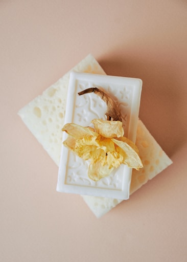 A decorative white soap with embossed patterns sits on a natural sponge, topped by a dried, pale yellow flower. The background consists of a light beige surface.