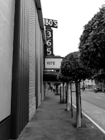 A sidewalk runs alongside a building with a large vertical sign displaying 'BO'S 365'. The marquee reads 'VOTE'. Trees with trimmed foliage line the sidewalk, and a pole with a parking sign stands nearby. The scene is in black and white, indicating an urban setting with no visible pedestrians.