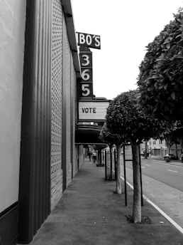 A sidewalk runs alongside a building with a large vertical sign displaying 'BO'S 365'. The marquee reads 'VOTE'. Trees with trimmed foliage line the sidewalk, and a pole with a parking sign stands nearby. The scene is in black and white, indicating an urban setting with no visible pedestrians.