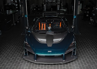 A sleek, high-performance sports car with a dark teal exterior and orange accents is parked in a garage. The car features an aerodynamic design with a prominent black front hood. The garage setting includes tile flooring and appears to have various tools and equipment in the background.