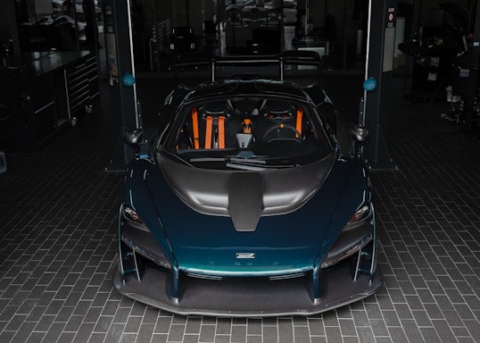 A sleek, high-performance sports car with a dark teal exterior and orange accents is parked in a garage. The car features an aerodynamic design with a prominent black front hood. The garage setting includes tile flooring and appears to have various tools and equipment in the background.