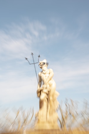 A statue of a classical figure holding a trident stands prominently, possibly portraying a mythological god. The image is slightly blurred, adding a sense of motion or mystical quality. Surrounding the statue is a fountain with water spraying upwards. The sky is a bright blue, contrasting with the pale stone color of the statue.