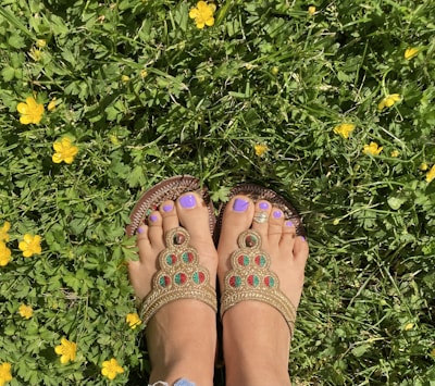Feet with purple-painted toenails are wearing ornate sandals on grass surrounded by small yellow flowers.