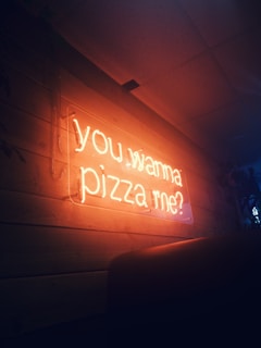 A glowing neon sign displays the words 'you wanna pizza me?' against a wooden wall. The warm orange light contrasts with the dark surroundings, creating a cozy, inviting atmosphere.