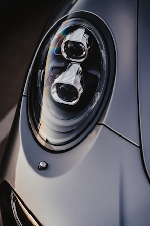 A close-up of a sleek car headlight with a modern and aerodynamic design, featuring intricate details and reflective surfaces. The lighting highlights the smooth contours and craftsmanship.