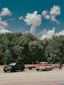 Three vehicles are parked on a paved area in front of a dense forest under a sky with scattered clouds. The first vehicle from the left is a black SUV, the second is a vintage red and white pickup truck towing a trailer, and the third is a dark-colored pickup truck also towing a trailer.