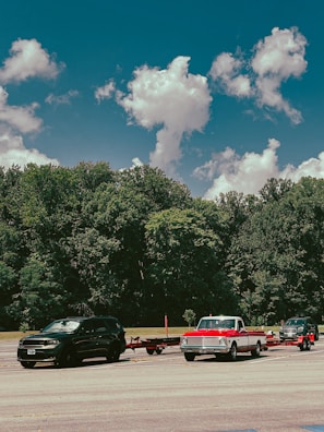 Three vehicles are parked on a paved area in front of a dense forest under a sky with scattered clouds. The first vehicle from the left is a black SUV, the second is a vintage red and white pickup truck towing a trailer, and the third is a dark-colored pickup truck also towing a trailer.