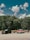 Three vehicles are parked on a paved area in front of a dense forest under a sky with scattered clouds. The first vehicle from the left is a black SUV, the second is a vintage red and white pickup truck towing a trailer, and the third is a dark-colored pickup truck also towing a trailer.