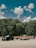 Three vehicles are parked on a paved area in front of a dense forest under a sky with scattered clouds. The first vehicle from the left is a black SUV, the second is a vintage red and white pickup truck towing a trailer, and the third is a dark-colored pickup truck also towing a trailer.