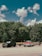 Three vehicles are parked on a paved area in front of a dense forest under a sky with scattered clouds. The first vehicle from the left is a black SUV, the second is a vintage red and white pickup truck towing a trailer, and the third is a dark-colored pickup truck also towing a trailer.