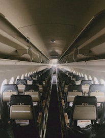 An empty airplane cabin with rows of seats on either side of a central aisle. Overhead compartments are closed, and the lighting is dim, creating a quiet and serene atmosphere. The seats have dark covers, and the windows lining the cabin allow some natural light to fill the space.