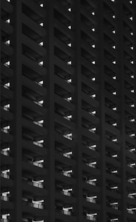A black and white photo of a high-rise building with repetitive horizontal lines. The structure appears to be a series of balconies or windows, emphasizing geometric patterns and symmetry. The contrast between the dark areas and the illuminated sections creates a striking visual effect.