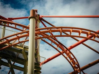 A complex network of roller coaster tracks intertwines against a partially cloudy blue sky. The tracks are predominantly orange and brown, with vertical support beams adding to the structural intricacies.