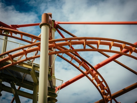 A complex network of roller coaster tracks intertwines against a partially cloudy blue sky. The tracks are predominantly orange and brown, with vertical support beams adding to the structural intricacies.