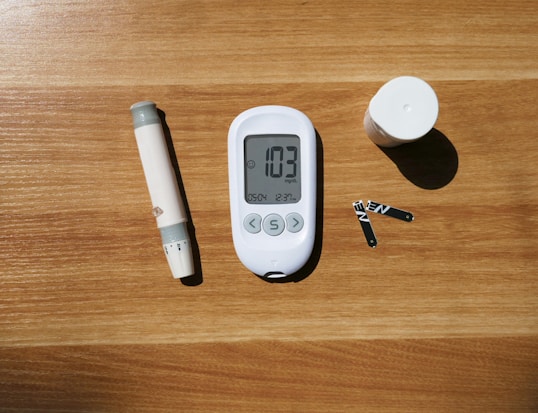 A glucose meter displaying a reading of 103 mg/dL is placed on a wooden surface. Beside it are a lancing device, test strips, and a small cylindrical container, likely used for holding more strips.