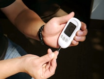 A person is holding a white glucose meter in their hands, preparing to take a blood sugar test. The focus is on the meter and the person's hands, with sunlight casting shadows across the scene.