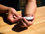 A person is using a white device, possibly a blood glucose meter, on a wooden table. The person is holding the device in one hand while the other hand seems to be touching the tip of a finger, likely for blood sampling.