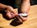 A person is using a white device, possibly a blood glucose meter, on a wooden table. The person is holding the device in one hand while the other hand seems to be touching the tip of a finger, likely for blood sampling.