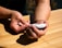 A person is using a white device, possibly a blood glucose meter, on a wooden table. The person is holding the device in one hand while the other hand seems to be touching the tip of a finger, likely for blood sampling.
