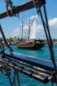 A wooden sailboat with a large white sail is on the ocean, seen through rigging in the foreground. The boat has several people on board and a banner that reads 'Jolly Pirates.' The water is a deep turquoise, and there is a coastline with palm trees in the background under a blue sky with some clouds.