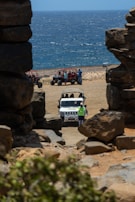 A rugged coastline with a view of the ocean, framed by large rock formations. In the foreground, a white jeep is parked on sandy terrain with several people around it. To the side, a group of off-road vehicles and people are visible on the beach. The scene conveys a sense of adventure and exploration.