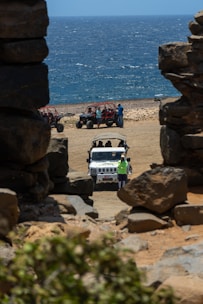 A rugged coastline with a view of the ocean, framed by large rock formations. In the foreground, a white jeep is parked on sandy terrain with several people around it. To the side, a group of off-road vehicles and people are visible on the beach. The scene conveys a sense of adventure and exploration.