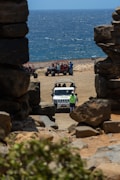 A rugged coastline with a view of the ocean, framed by large rock formations. In the foreground, a white jeep is parked on sandy terrain with several people around it. To the side, a group of off-road vehicles and people are visible on the beach. The scene conveys a sense of adventure and exploration.