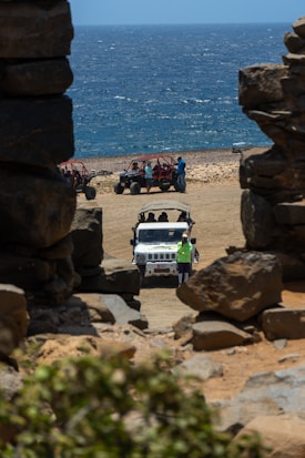 A rugged coastline with a view of the ocean, framed by large rock formations. In the foreground, a white jeep is parked on sandy terrain with several people around it. To the side, a group of off-road vehicles and people are visible on the beach. The scene conveys a sense of adventure and exploration.