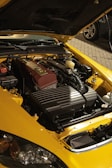 A yellow car with its hood open, revealing a clean and organized engine. The engine components are neatly arranged, with a prominent maroon valve cover. The car is parked on a cobblestone driveway, and part of another vehicle is visible in the background.