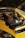 A yellow car with its hood open, revealing a clean and organized engine. The engine components are neatly arranged, with a prominent maroon valve cover. The car is parked on a cobblestone driveway, and part of another vehicle is visible in the background.