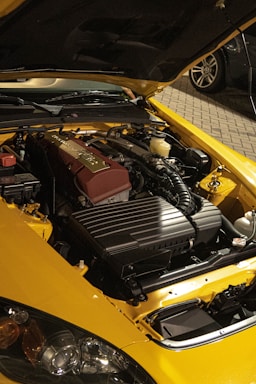 A yellow car with its hood open, revealing a clean and organized engine. The engine components are neatly arranged, with a prominent maroon valve cover. The car is parked on a cobblestone driveway, and part of another vehicle is visible in the background.