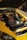 A yellow car with its hood open, revealing a clean and organized engine. The engine components are neatly arranged, with a prominent maroon valve cover. The car is parked on a cobblestone driveway, and part of another vehicle is visible in the background.