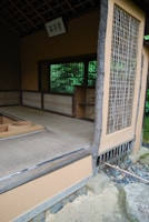 An open traditional Japanese tea room with tatami mat flooring and a rustic wooden structure. The space includes sliding shoji-style windows and subtle earthy colors, surrounded by natural greenery. A wooden sign with calligraphy hangs above the entrance.
