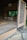 An open traditional Japanese tea room with tatami mat flooring and a rustic wooden structure. The space includes sliding shoji-style windows and subtle earthy colors, surrounded by natural greenery. A wooden sign with calligraphy hangs above the entrance.
