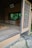 An open traditional Japanese tea room with tatami mat flooring and a rustic wooden structure. The space includes sliding shoji-style windows and subtle earthy colors, surrounded by natural greenery. A wooden sign with calligraphy hangs above the entrance.