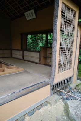 An open traditional Japanese tea room with tatami mat flooring and a rustic wooden structure. The space includes sliding shoji-style windows and subtle earthy colors, surrounded by natural greenery. A wooden sign with calligraphy hangs above the entrance.