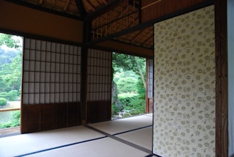 An interior of a traditional Japanese room featuring tatami mats on the floor, sliding shoji doors, and a view of lush green vegetation through an open doorway. The room is decorated with wooden beams and a floral-patterned panel.