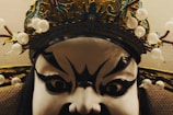 A close-up of a theatrical mask featuring exaggerated black and white facial paint with bold, expressive eyes. The mask is adorned with an elaborate, ornate headpiece decorated with white beads and intricate detailing.