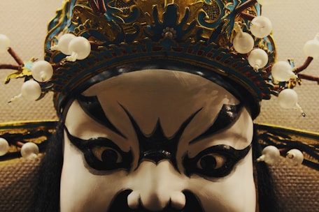 A close-up of a theatrical mask featuring exaggerated black and white facial paint with bold, expressive eyes. The mask is adorned with an elaborate, ornate headpiece decorated with white beads and intricate detailing.