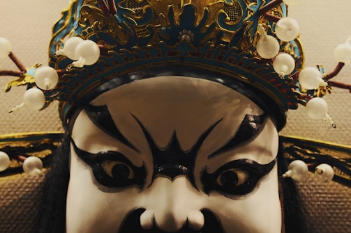 A close-up of a theatrical mask featuring exaggerated black and white facial paint with bold, expressive eyes. The mask is adorned with an elaborate, ornate headpiece decorated with white beads and intricate detailing.