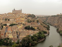 A historic town with dense architecture featuring a variety of buildings surrounded by a winding river. The large castle-like structure dominates the skyline, set against a backdrop of rolling hills and greenery.