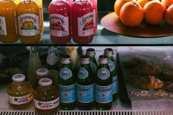 A display shelf featuring bottles of various drinks such as juices and sparkling water. Bundaberg bottles are at the top with flavors like tropical mango and pink grapefruit. Next to them are fresh oranges on a platter. Below, Pellegrino sparkling water bottles and small round bottles of Martinelli's apple juice are arranged. To the right, several croissants rest on a lined tray.