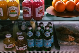 A display shelf featuring bottles of various drinks such as juices and sparkling water. Bundaberg bottles are at the top with flavors like tropical mango and pink grapefruit. Next to them are fresh oranges on a platter. Below, Pellegrino sparkling water bottles and small round bottles of Martinelli's apple juice are arranged. To the right, several croissants rest on a lined tray.
