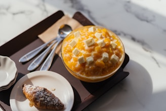 A delicious-looking dessert featuring a bowl of mango shaved ice topped with chunks of cheesecake sits on a dark serving tray. Next to it, a small plate holds an almond croissant. Several silver spoons rest on a beige napkin beside the bowl, all placed on a marble surface.